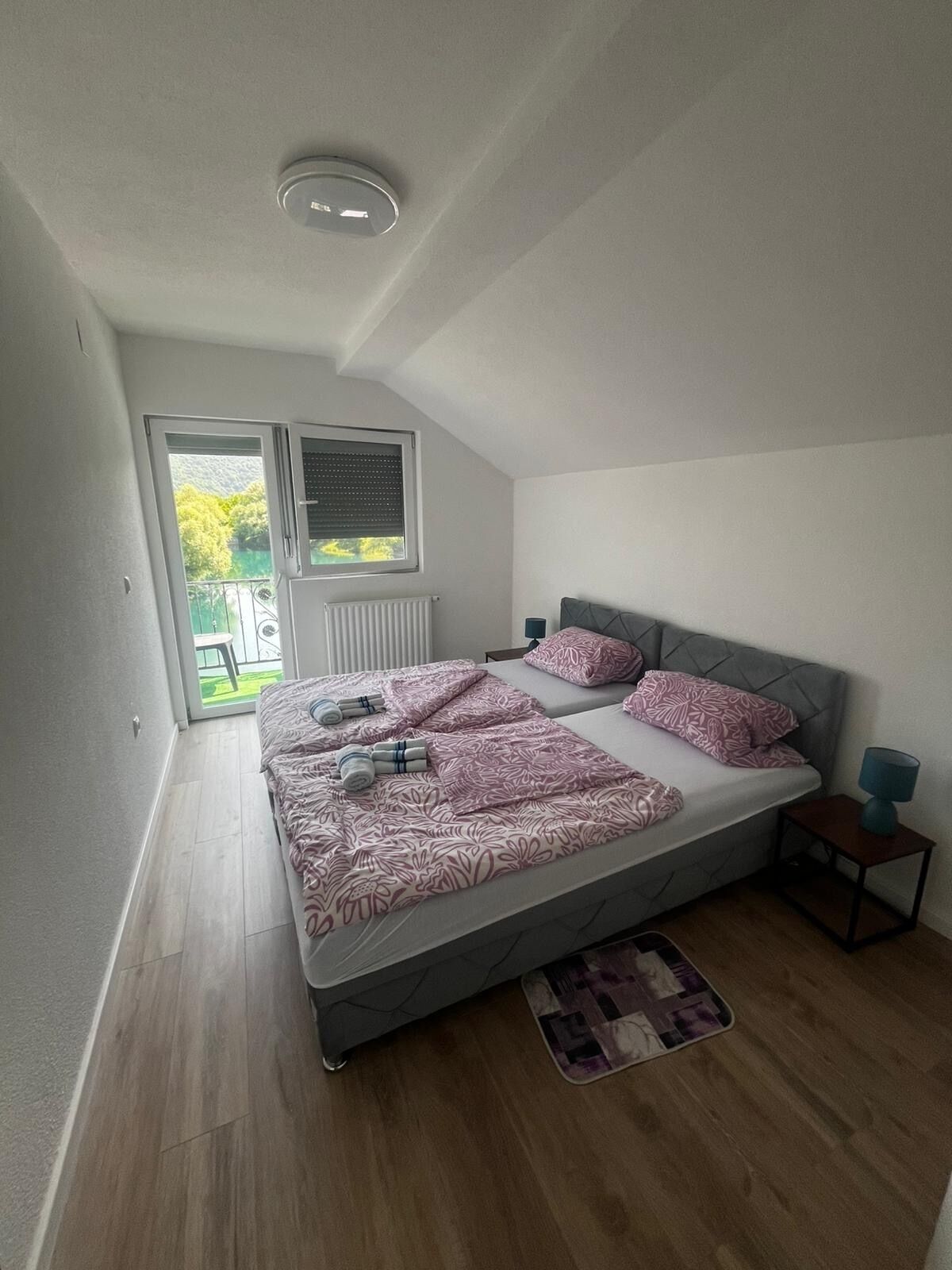 Une chambre à coucher comprenant un lit, une table de chevet, une lampe, une fenêtre avec des stores et une vue sur la verdure à l’extérieur.