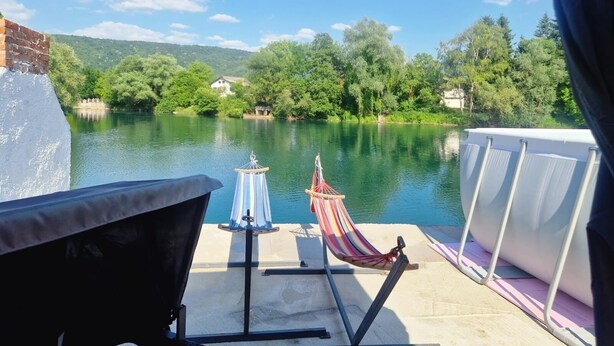 Un balcon avec un hamac, un barbecue et une vue sur un lac, avec des arbres et des maisons en arrière-plan.