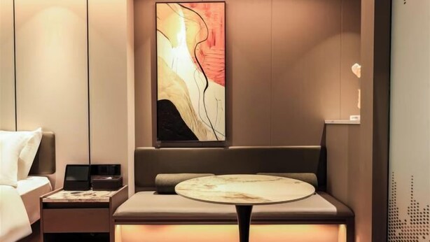 Ein modernes Hotelzimmer mit einem Bett, einem Nachttisch mit Lampe, einer Sitzecke mit gepolstertem Sitz und einem an der Wand hängenden abstrakten Kunstwerk.