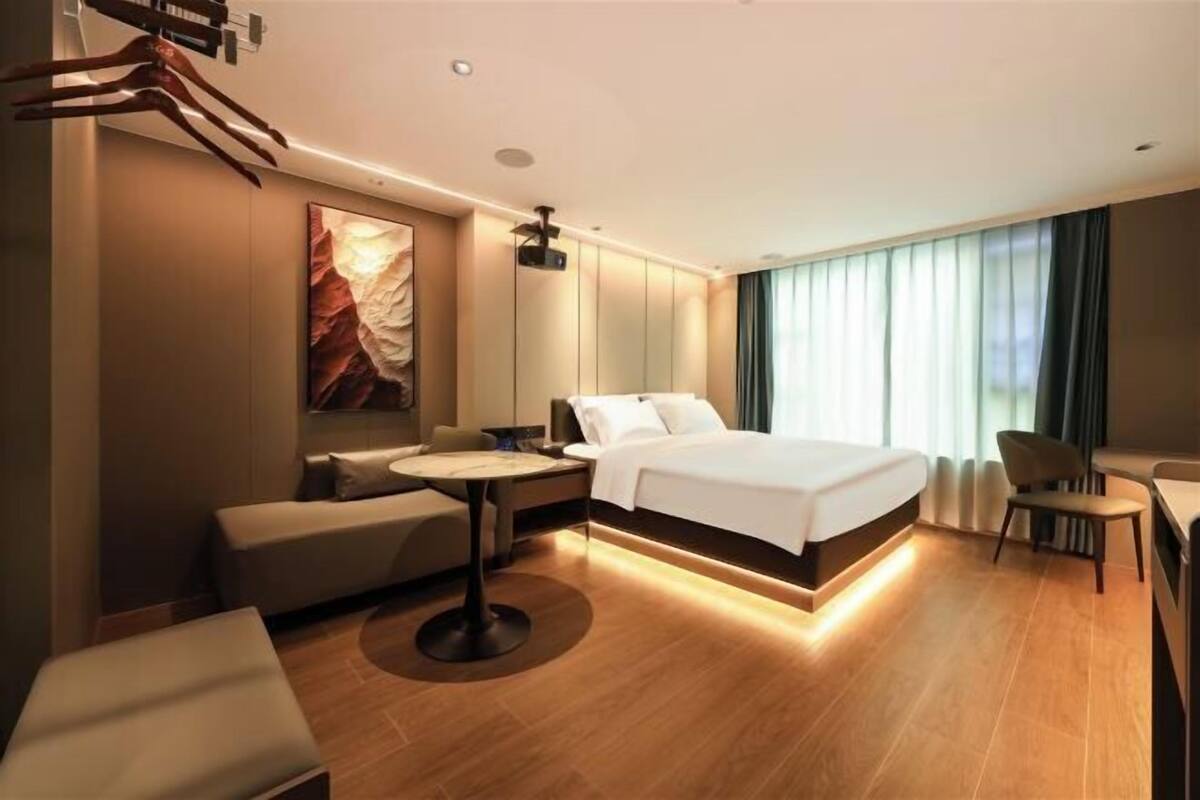 Una habitación de hotel moderna con una cama grande, una zona de estar con mesa y un amplio ventanal con cortinas.