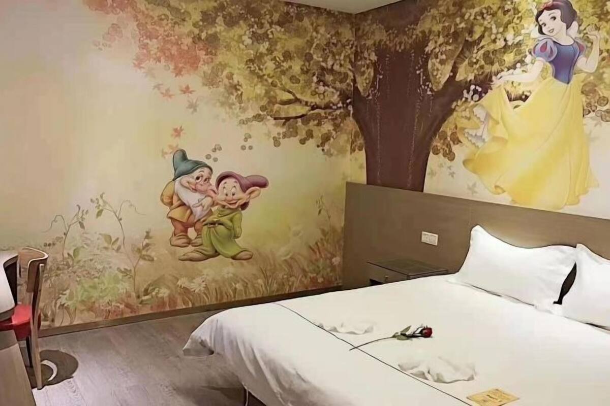 Une chambre d’hôtel avec une fresque murale représentant Blanche-Neige et les Sept Nains, un lit avec une literie blanche et une table de chevet avec une lampe.