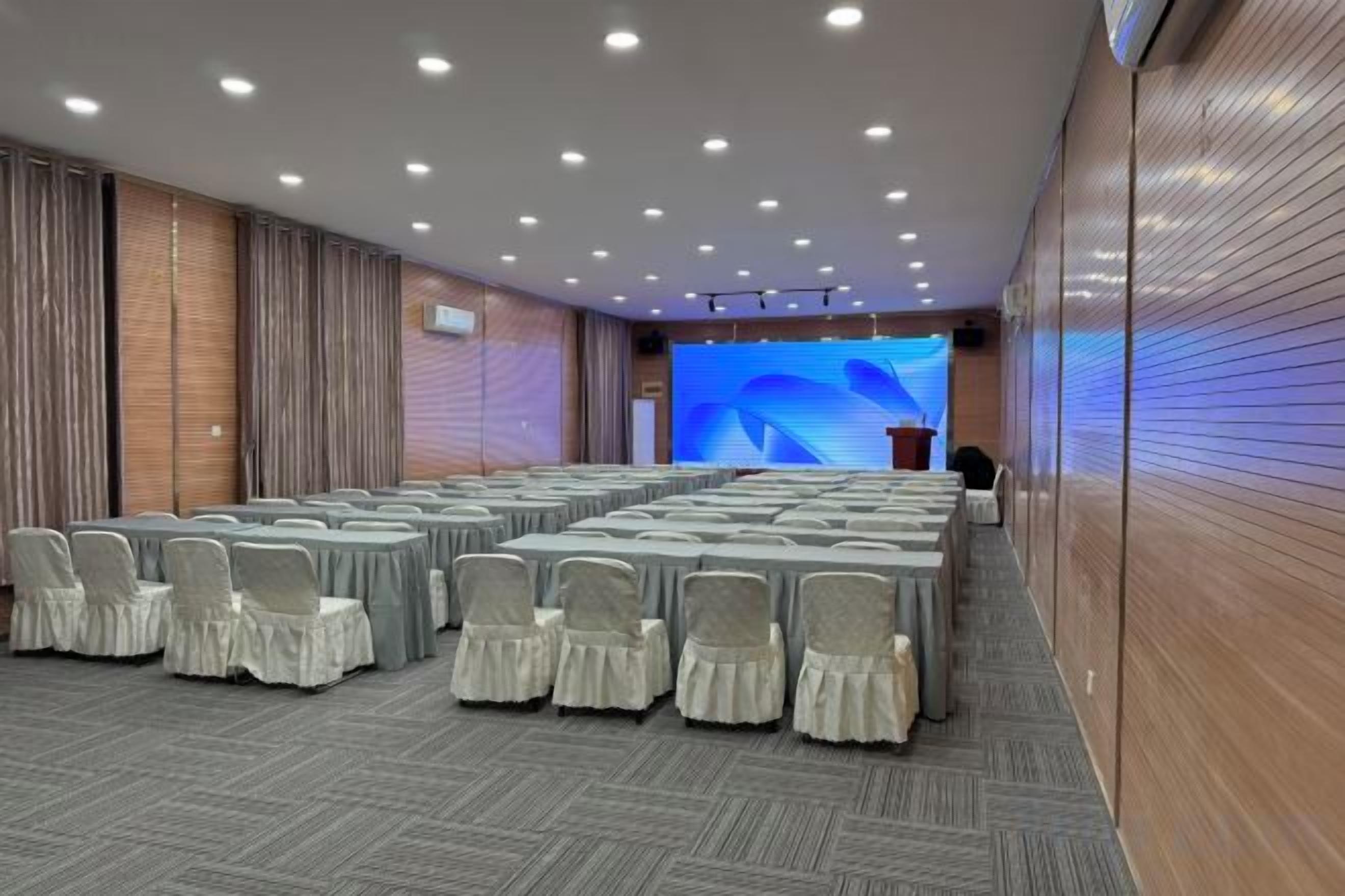 A conference room with rows of tables and chairs, a stage with a large screen, and a podium.