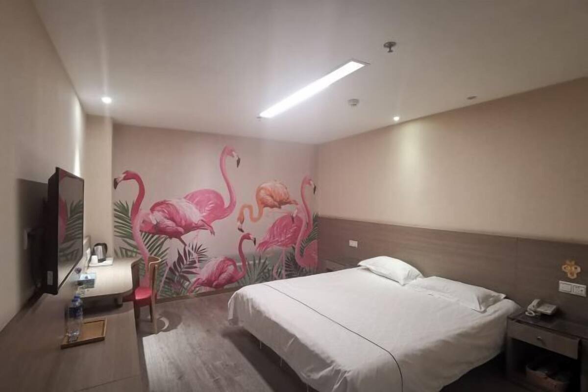 Une chambre d’hôtel comprenant un lit, une table de chevet, un bureau avec une lampe, un miroir et un mur orné d’une fresque représentant des flamants roses.
