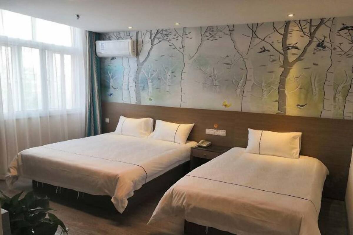 Une chambre d’hôtel avec deux lits, une grande fenêtre avec des rideaux légers, un climatiseur et une fresque murale représentant des arbres et des oiseaux.