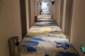 Un couloir d’hôtel avec un tapis à motifs, une poubelle et un écran numérique.
