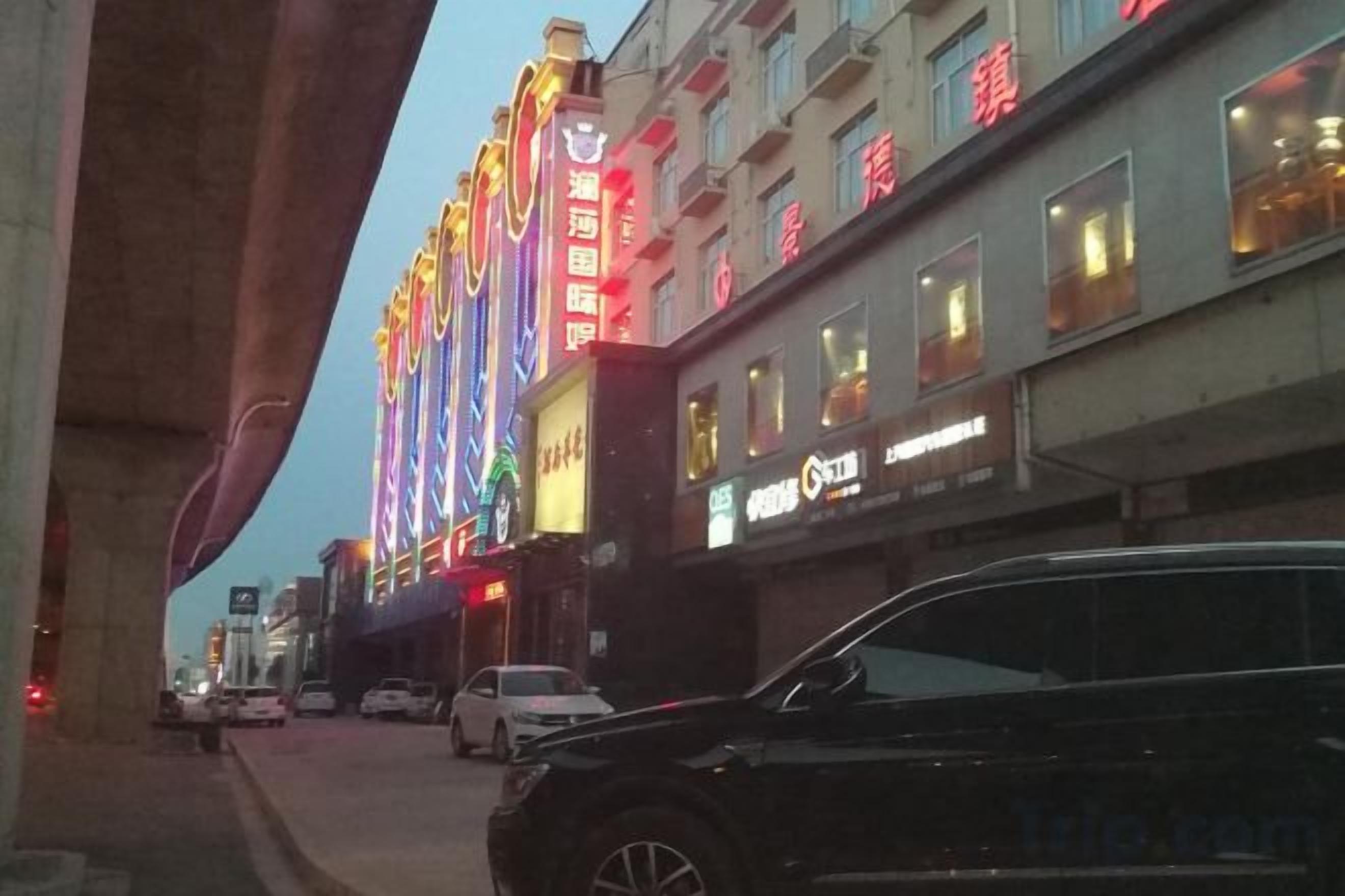 A street scene with a brightly lit building, cars parked along the side, and a pedestrian walkway.