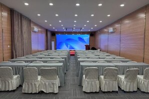 Ein Konferenzraum mit Tischen und Stühlen in Reihen, einer großen Leinwand im Vordergrund und einer Wand mit Holzpaneelen.