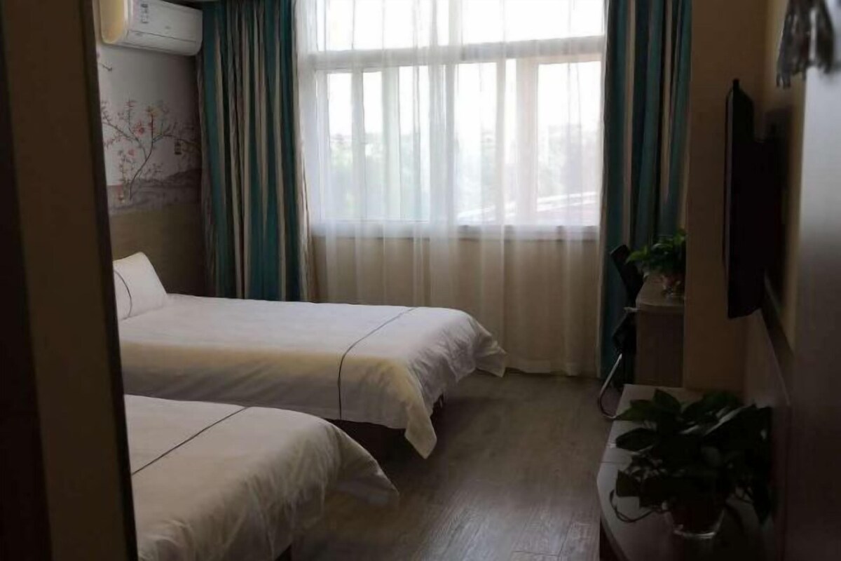 Une chambre d’hôtel avec deux lits, une fenêtre avec des rideaux légers, un climatiseur fixé au mur et une plante en pot sur une table d’appoint.
