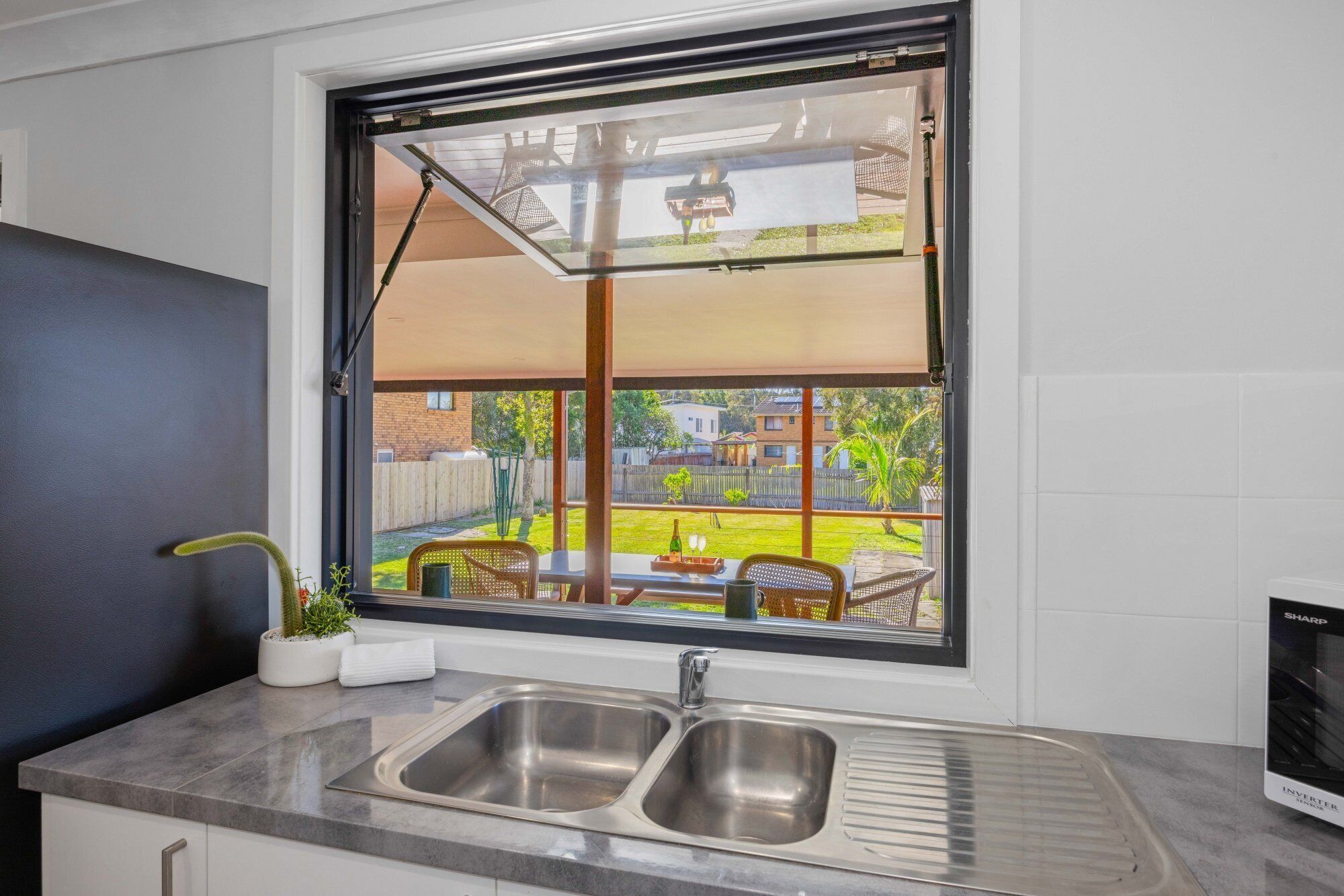 A kitchen with a large window, two stainless steel sinks, and a view of a backyard with outdoor furniture.