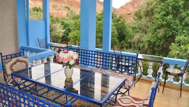 A blue-tiled balcony with a glass table and chairs, a vase of flowers, and a view of green trees and red hills. - A Secret Garden (Toudgha El Oulia)