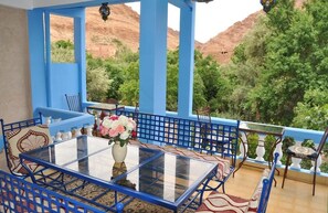 Un balcón con baldosas azules, una mesa y sillas de vidrio, un jarrón con flores y vistas a árboles verdes y colinas rojas.