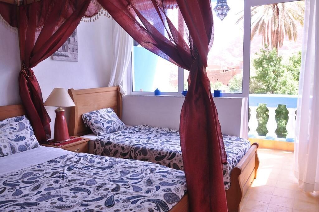 Une chambre avec deux lits, une fenêtre donnant sur des palmiers et un balcon avec des balustrades bleues.