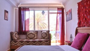 A bedroom with a large bed, a sofa with patterned cushions, a window with curtains, and a view of palm trees. - A Secret Garden (Toudgha El Oulia)