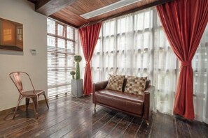 A room with a leather sofa, a wooden chair, a potted plant, and a large window with red curtains.