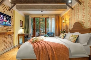 A bedroom with a large bed, a TV mounted on the wall, a window with curtains, and a wooden ceiling.