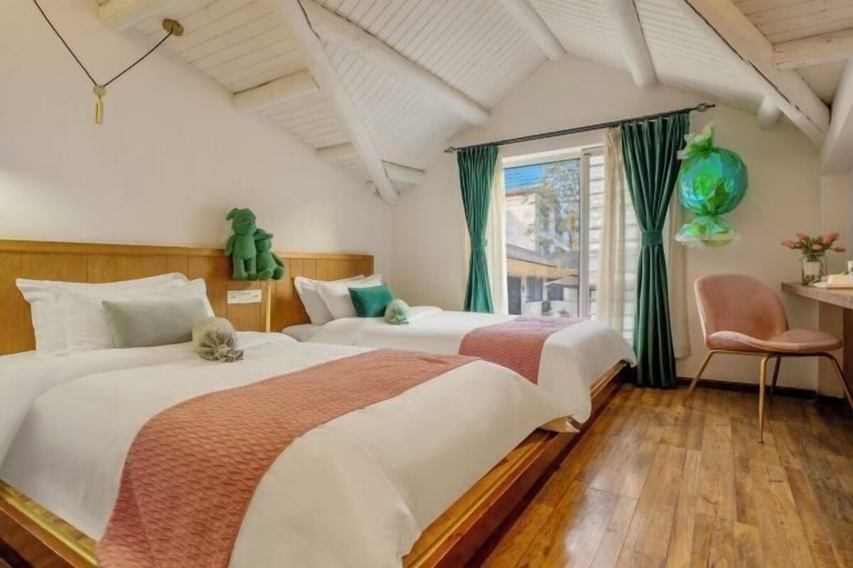Une chambre d’hôtel avec deux lits, un plancher en bois, une tête de lit en bois, une petite table avec une chaise et une fenêtre avec des rideaux.