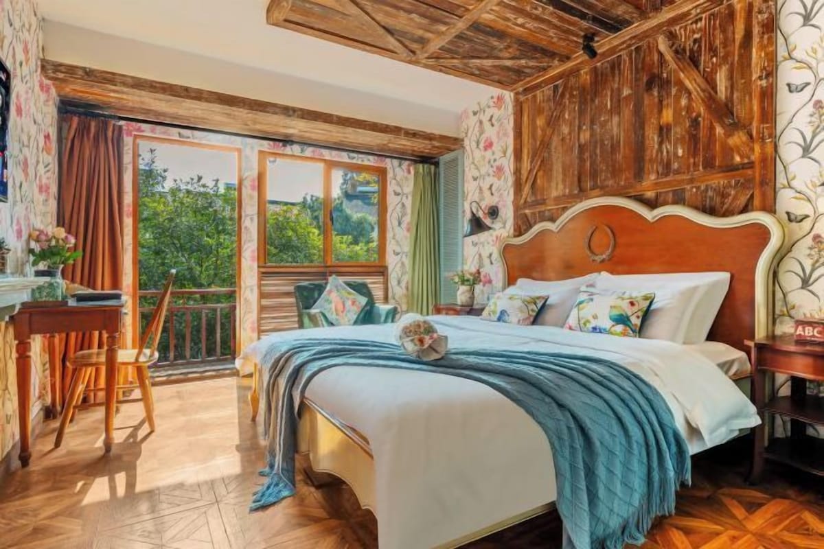 Une chambre à coucher avec une tête de lit en bois, un grand lit avec de nombreux oreillers, une table de chevet et une vue sur la verdure par la fenêtre.