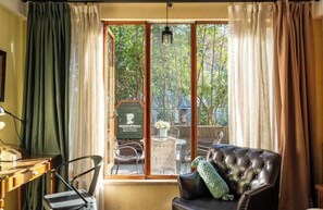 Une pièce dotée d’une grande porte-fenêtre coulissante qui donne sur un balcon offrant une vue sur les arbres et un jardin. On y trouve un canapé en cuir noir avec un coussin vert, une petite table et une chaise.