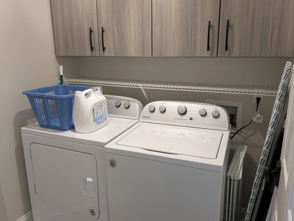 Un área de lavandería con lavadora y secadora de carga frontal, una cesta de ropa azul y una botella de detergente sobre la secadora.