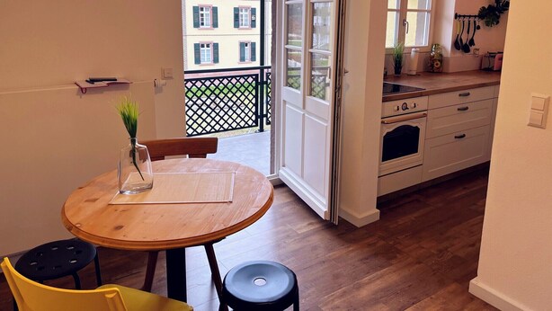 Een kleine keuken met een eethoek, een houten tafel en uitzicht op een gebouw door een geopende deur.