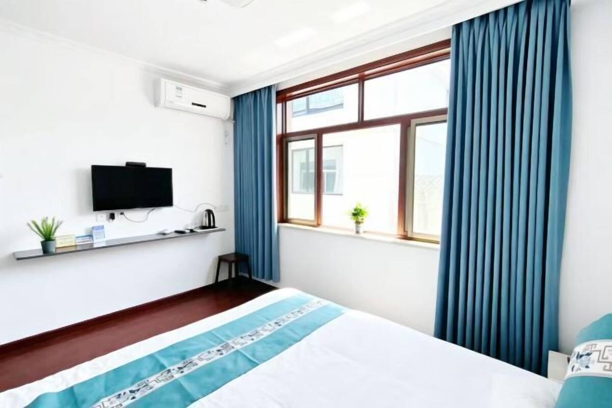 A hotel room with a bed, a TV mounted on the wall, a window with curtains, a small plant, and a shelf with items.