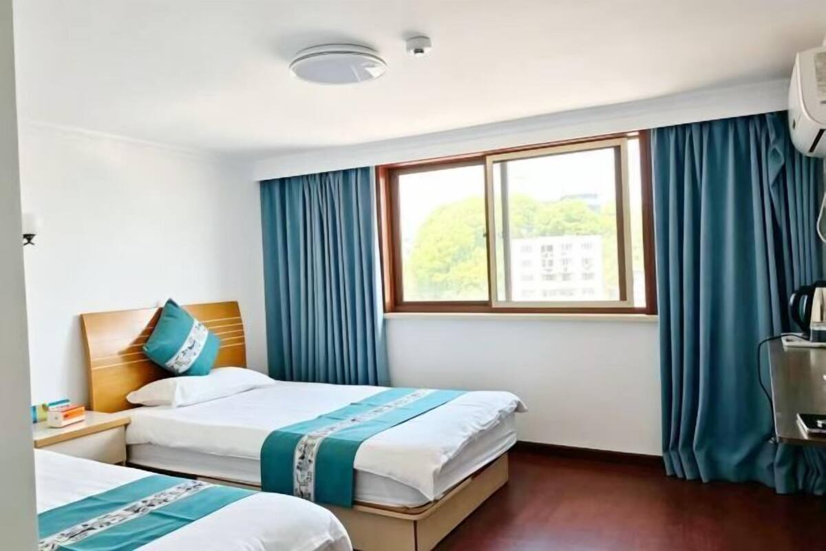A hotel room with two beds, a window with curtains, a ceiling light, and a wall-mounted air conditioner.