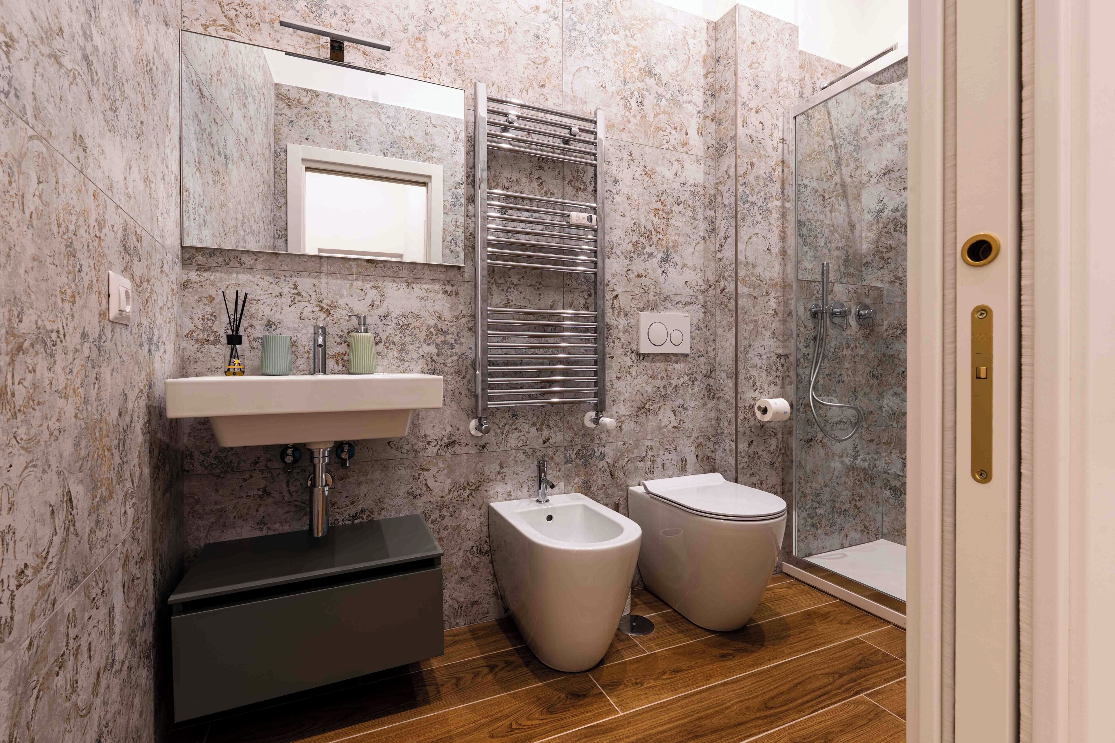 A bathroom with a marble wall, a sink, bidets, a shower, and a mirror.