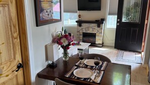 A dining area with a wooden table set for two, a vase of flowers, and a fireplace in the background.