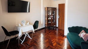 A living room with a flat-screen TV, a dining table with chairs, a green sofa, and a wooden cabinet. - B&B La Piazzetta (Pianillo)