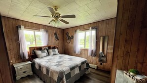 Een slaapkamer met een wand van houten panelen, een bed met quilt, een nachtkastje, een plafondventilator en een raam met gordijnen.