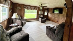 Een ruime woonkamer met een plafondventilator, een flatscreen-televisie aan de muur en een groot raam met uitzicht op de omgeving.