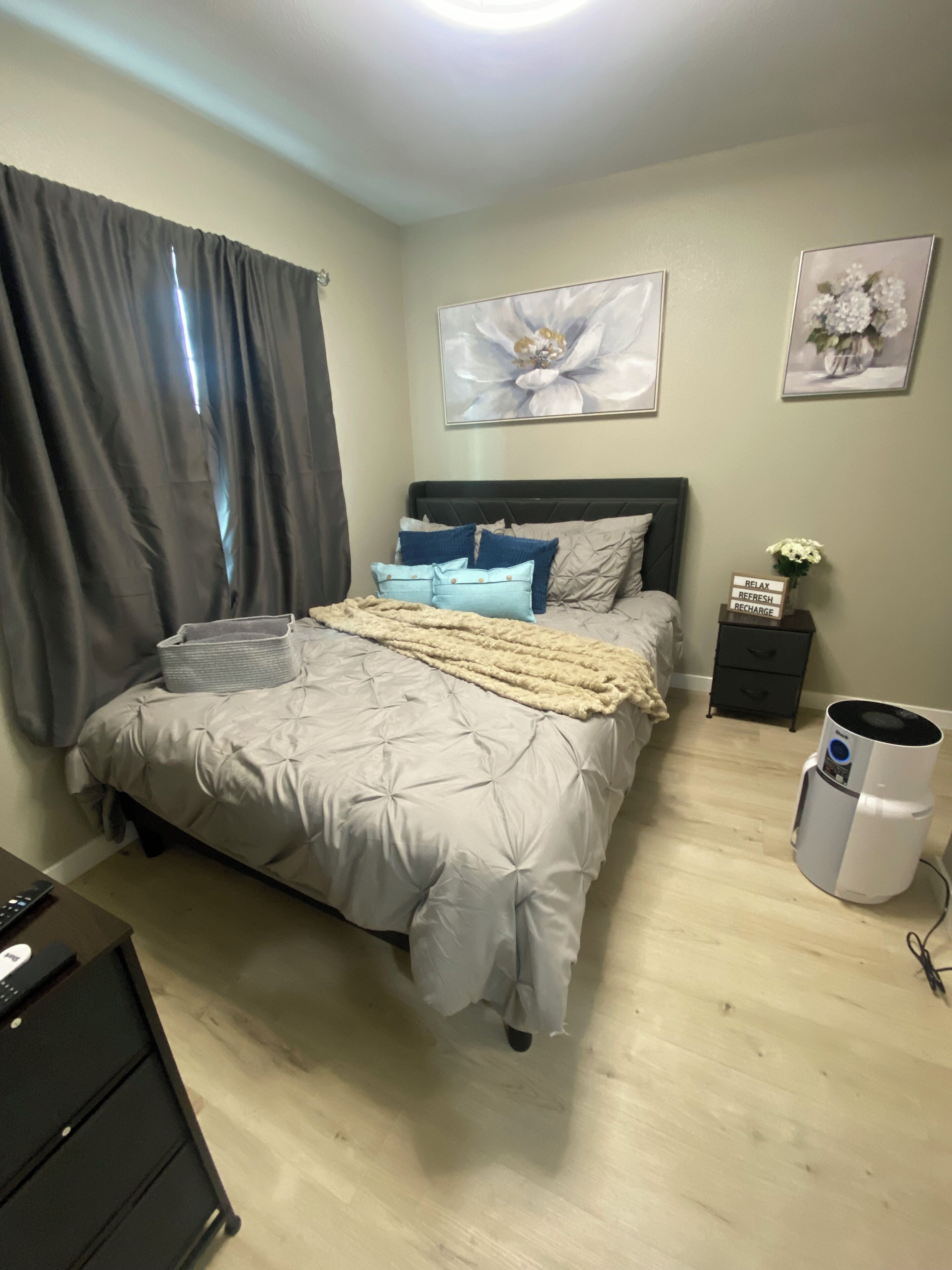 A bedroom with a bed, a bedside table, a nightstand, a trash can, and a wall-mounted artwork.