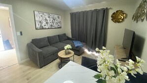 A modern living room with a grey sofa, a small round table with a vase of flowers, a flat-screen TV, and a large window with grey curtains. - Brand new 2-bedroom in charming Santa Maria with WiFi, AC (Santa Maria)