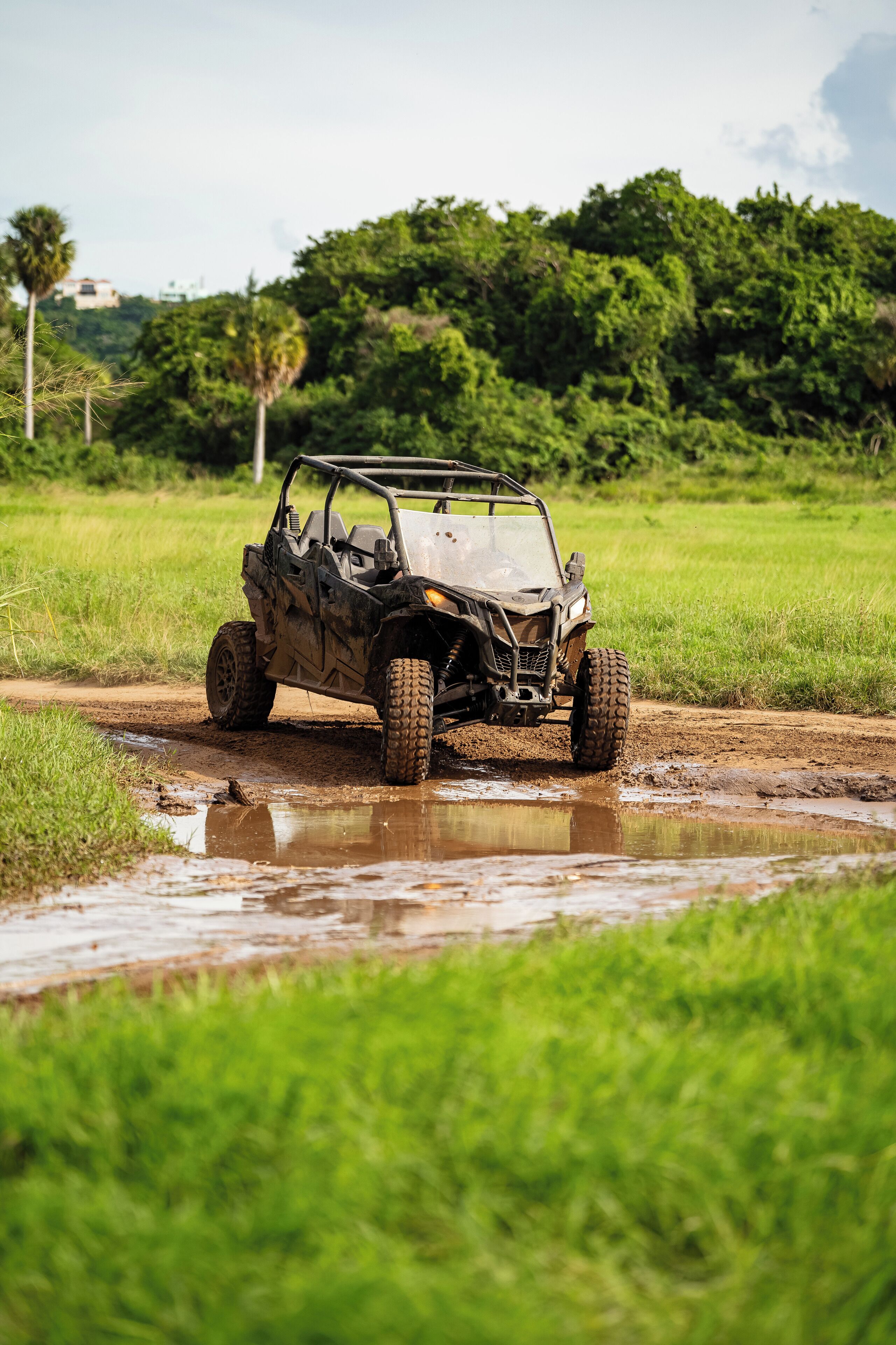 A vehicle navigating through a muddy puddle in a grassy field with trees in the background.