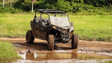 A vehicle navigating through a muddy puddle in a grassy field with trees in the background.