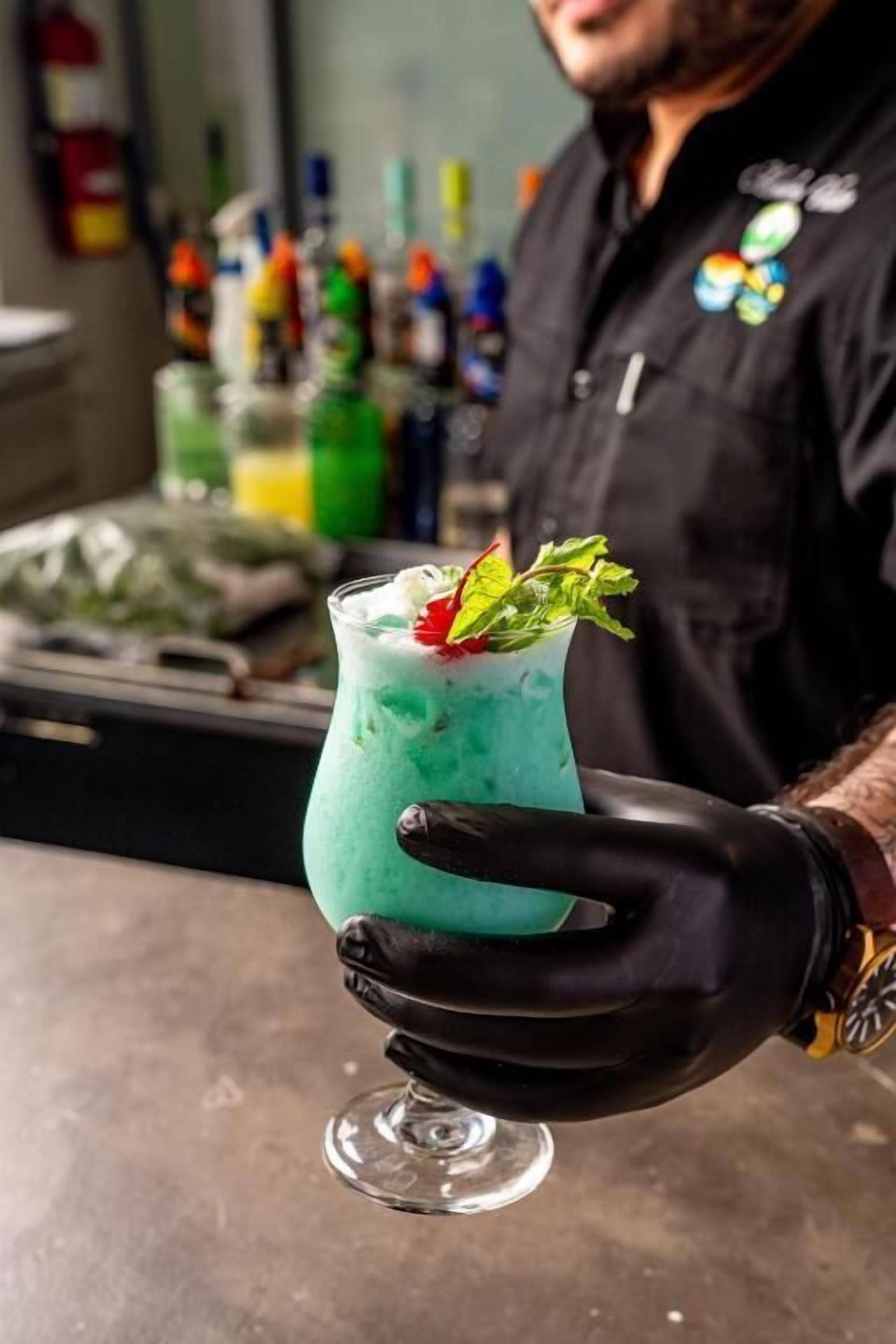 A bartender holding a green cocktail with a garnish, in a bar setting with various bottles in the background.
