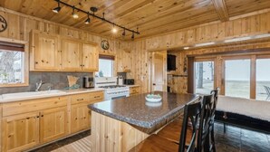 A wooden cabin interior with a kitchen, dining area, and large windows.