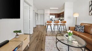 Un salon moderne avec un téléviseur à écran plat, un canapé en cuir marron, une table basse en verre et un coin repas avec des chaises, et une cuisine en arrière-plan.