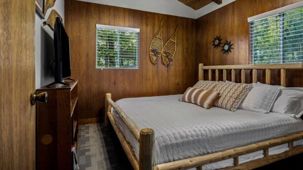 Une chambre avec des murs en bois, un lit agrémenté d’oreillers à motifs, une tête de lit en bois, une fenêtre avec des stores et une horloge fixée au mur.