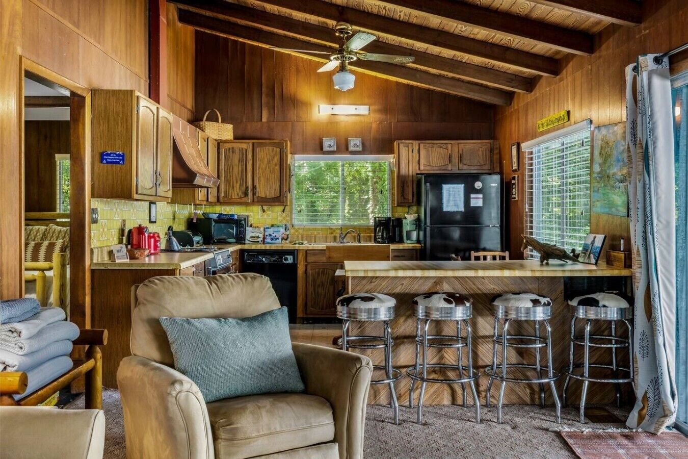Ein gemütliches Blockhaus-Interieur mit Holzausbau, einem Essbereich mit Hockern, einer Küche mit Geräten und einem Deckenventilator.
