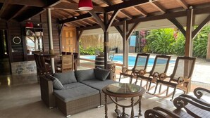 Un espace extérieur aménagé, abrité et doté de mobilier en osier, d’une table en verre et offrant une vue sur une piscine.