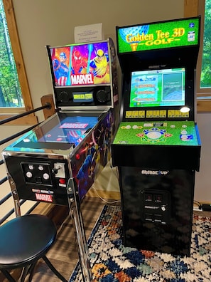 Two arcade machines with Marvel and Golden Tee 3D Golf games, a chair, and a window with a view of trees. - 
PERFECT HOME BASE FOR LAKES, TRAILS, SNOW SKIING! (Camp Connell)