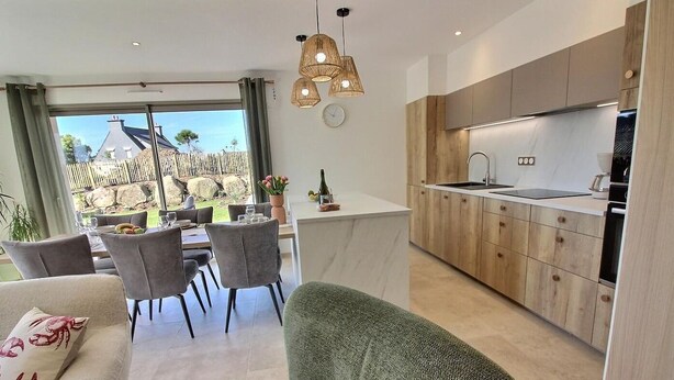 Una cocina moderna con armarios de madera, encimera blanca y horno empotrado. Hay una zona de comedor con una mesa y sillas, y se ve una casa a través de ventanas amplias.