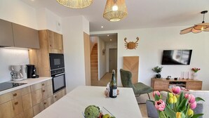 Cocina moderna con armarios de madera, horno empotrado y comedor con mesa blanca. Hay un televisor montado en la pared y una silla de mimbre en la sala de estar.