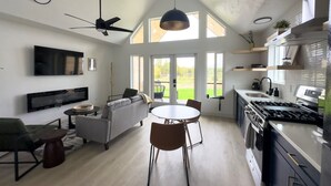 A modern kitchen with a built-in stove, a dining area with a round table and chairs, a sofa, and a TV mounted on the wall. - Lakefront Cottage with Sauna- #2 Coho (Wasilla)