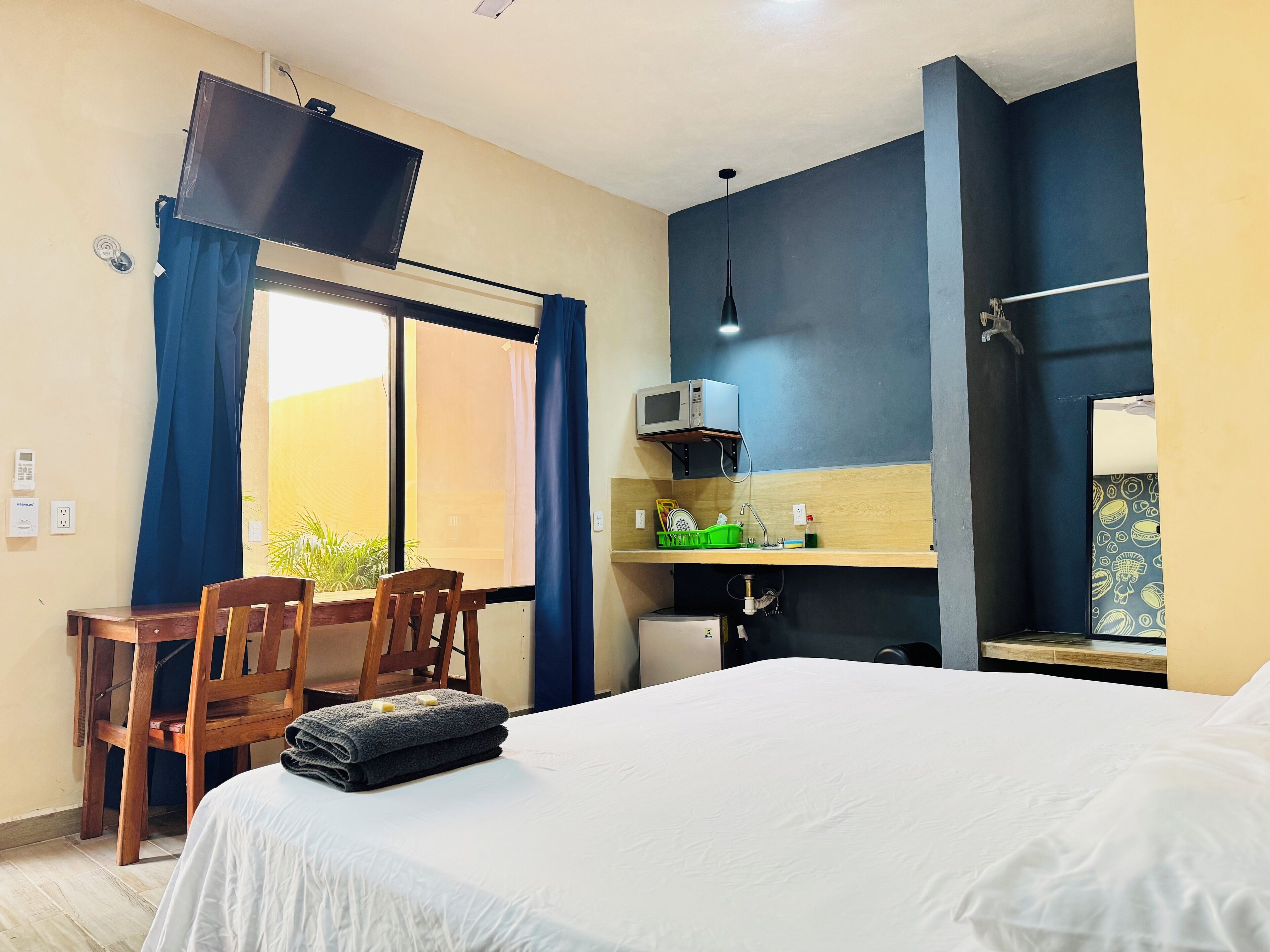 A hotel room with a bed, a desk with a chair, a window with curtains, a microwave, and a sink.