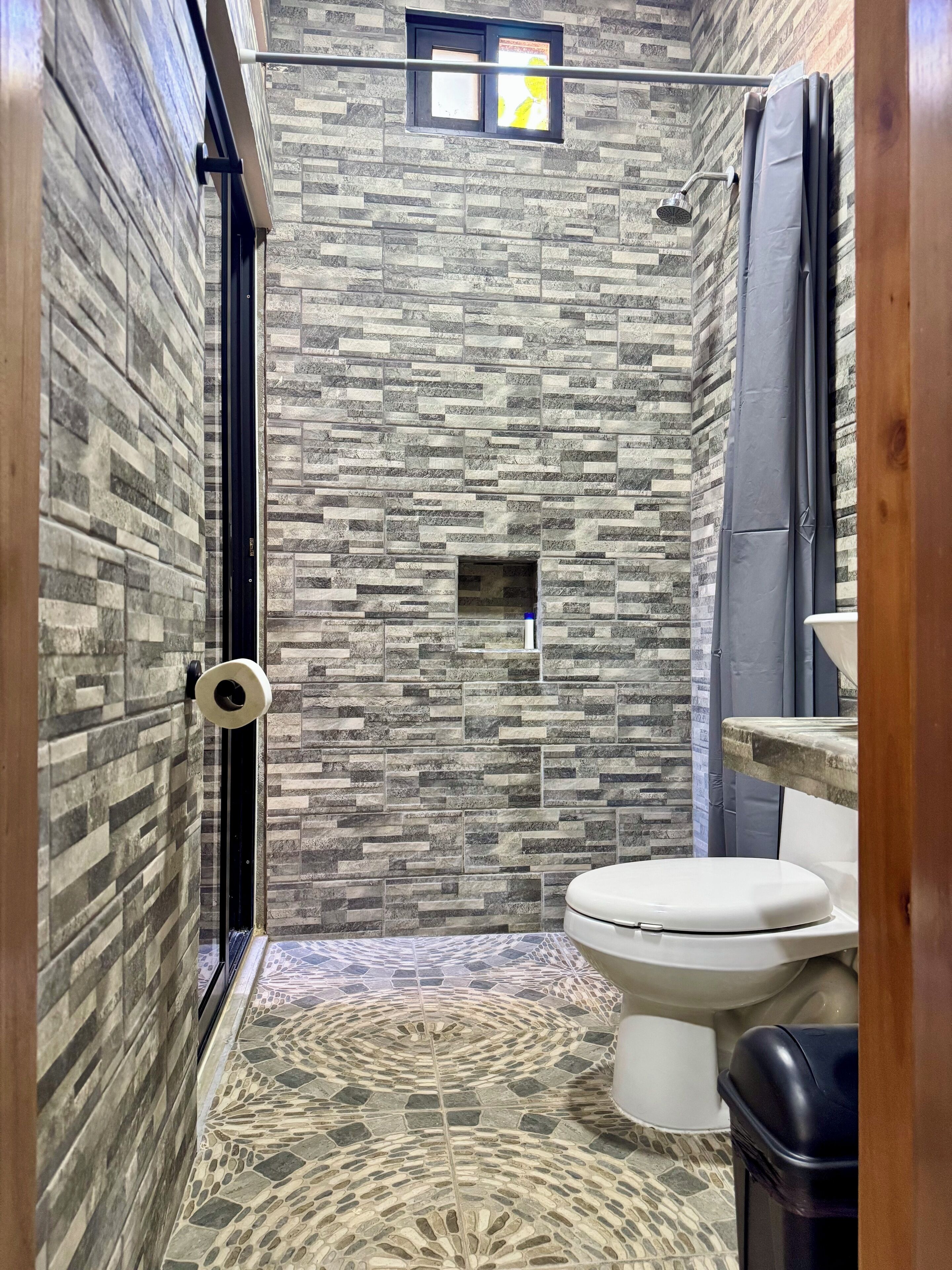 A bathroom with a stone-patterned shower wall, a toilet, and a trash bin.