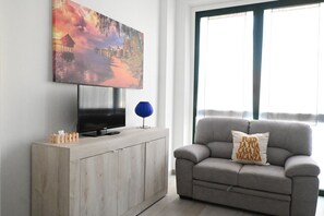Un salon avec un téléviseur posé sur un meuble en bois, une lampe bleue, un canapé gris avec un coussin et un grand tableau accroché au mur.