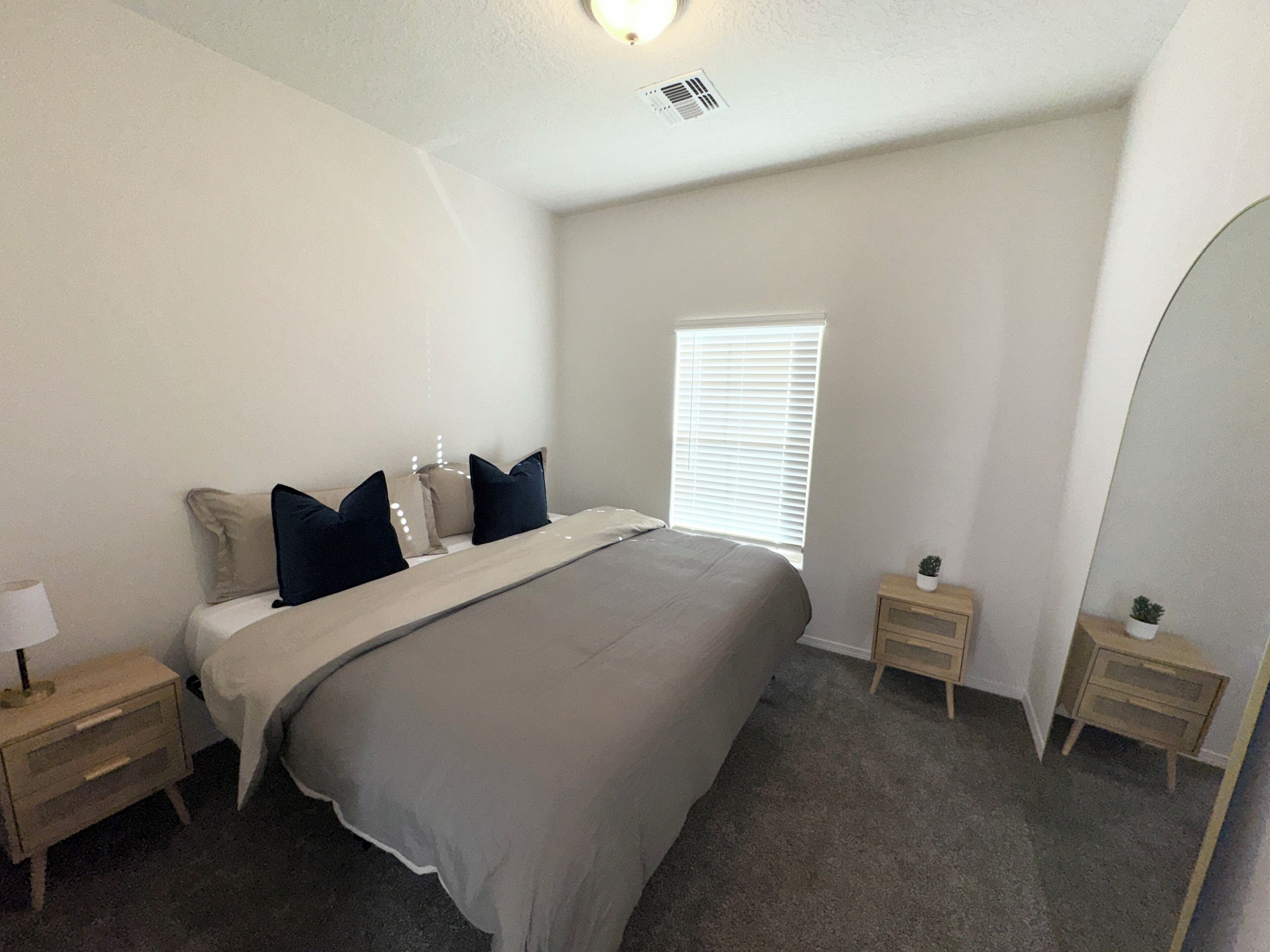 Une chambre à coucher comprenant un lit, deux tables de chevet, une commode, une fenêtre avec stores et un luminaire au plafond.