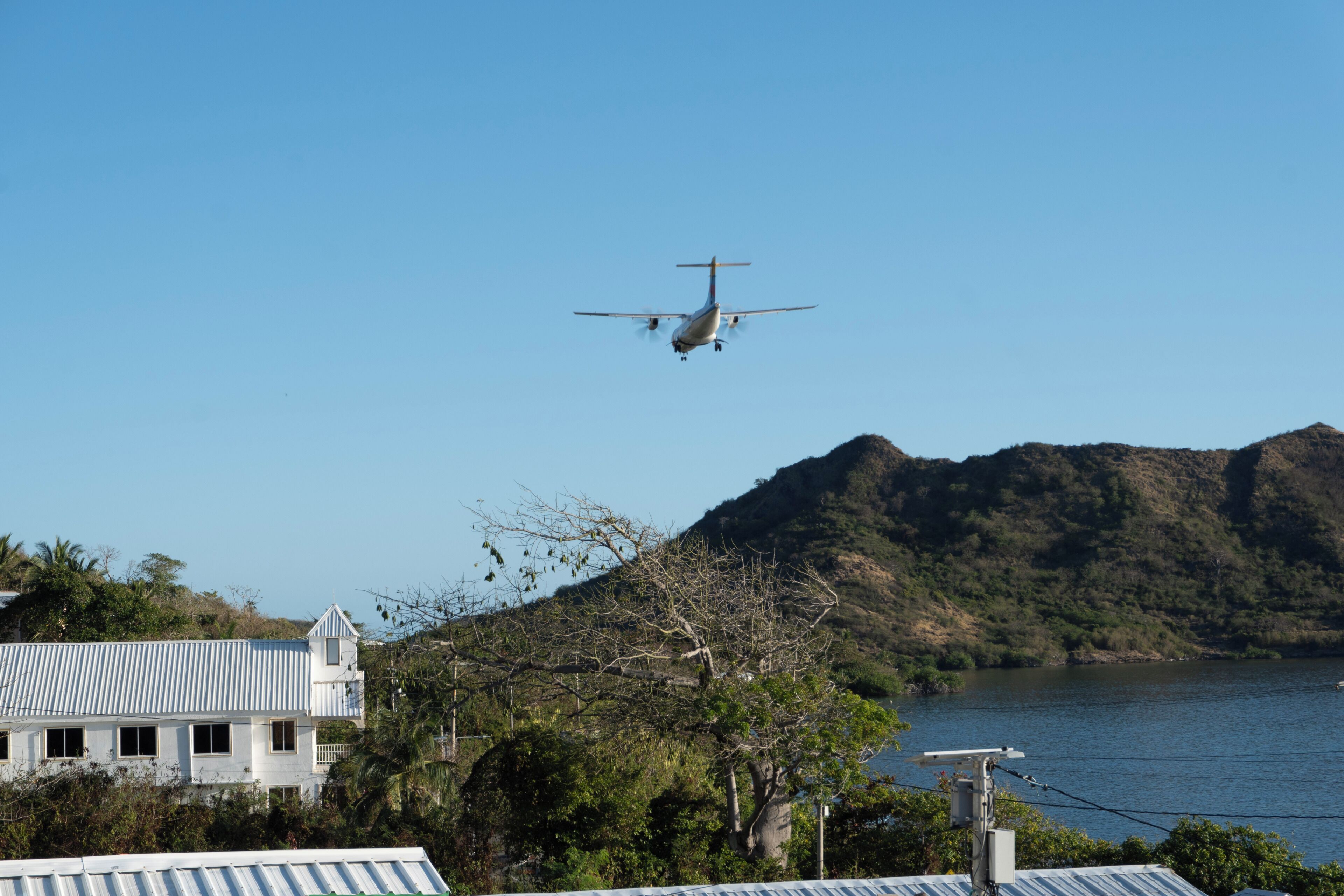A small aircraft flying over a coastal area with a house and hills in the background.